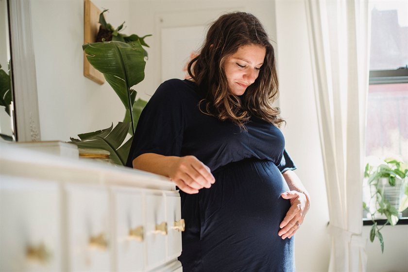 A pregnant woman standing in a sunlit bedroom, resting one hand on her belly.